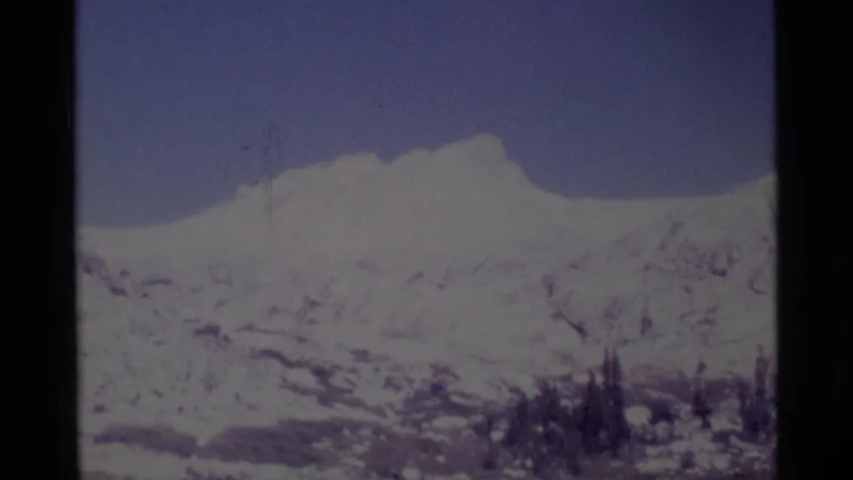 Faded 1975 8mm frame of the Minaret Wilderness with a snowy mountain ridge, exposed rock, blue sky, and dark evergreen cluster.