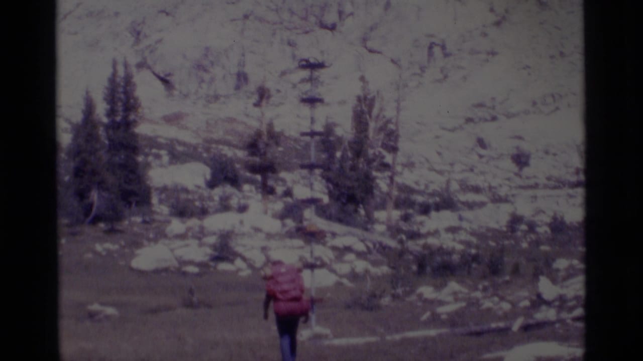 Hiker with a red backpack walking through snow and rocks below evergreen trees.