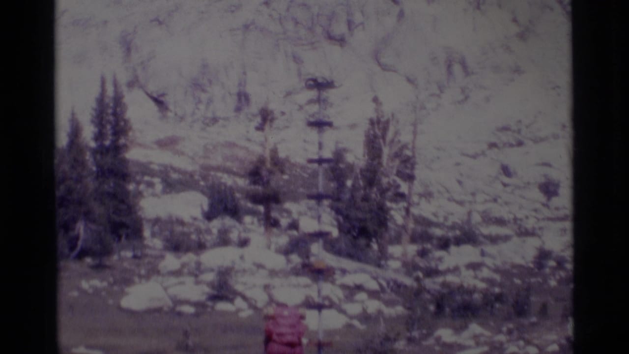Red-backpack hiker partly low in frame near snowy rocks and evergreen trees.