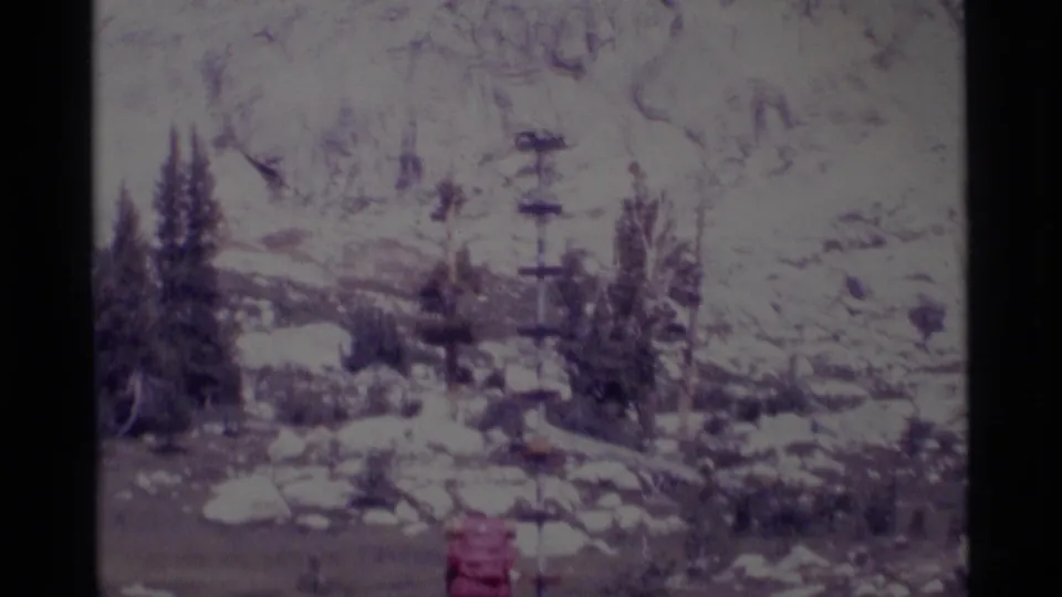 1975 California wilderness frame showing a red-backpack hiker crossing a snowy rocky area with trees and distant slopes.
