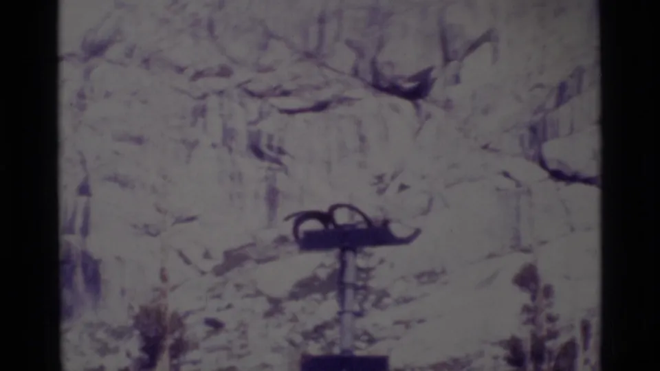 Close 1975 wilderness frame of a metal pole structure set before pale snowy rock and sparse trees.