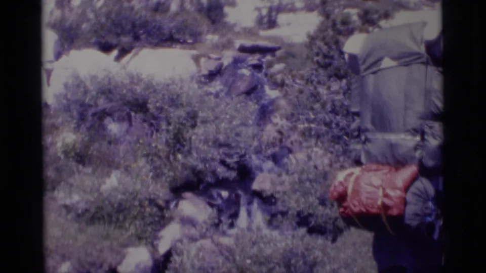 1975 Minaret Wilderness 8mm frame showing a hiker from behind with a red backpack near brush, boulders, and a small stream.