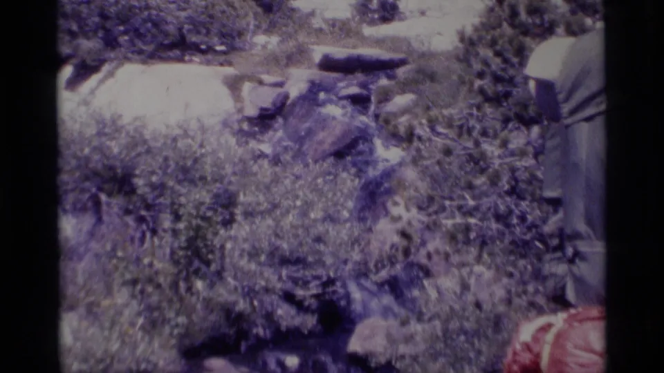 1975 Minaret Wilderness 8mm frame with brush, boulders, stream rocks, and the edge of a hiker carrying a red backpack.