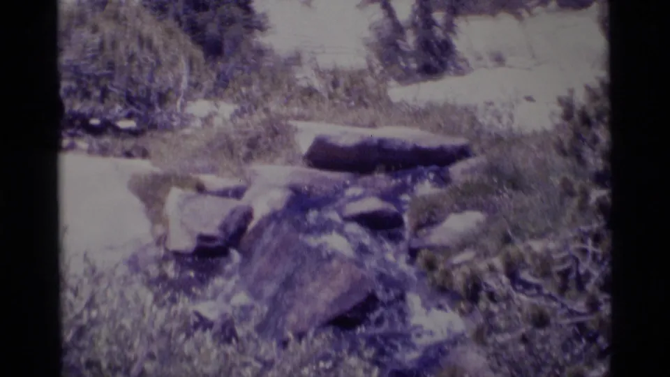 1975 Minaret Wilderness 8mm frame showing a rocky stream crossing surrounded by shrubs and boulders in bright sun.
