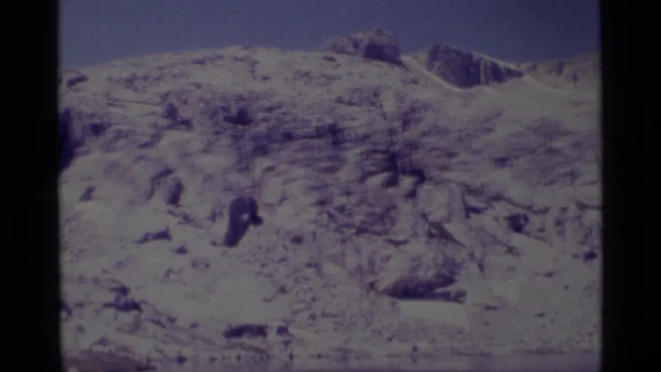 8mm home-movie frame of a snow-dusted alpine hillside above reservoir water, with clear sky and faded film color.