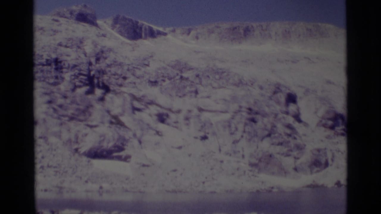 Rocky snowy mountainside with blue sky and a faint lake edge near the bottom.