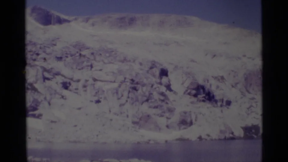 1975 8mm Minaret Wilderness frame showing a broad snow-dusted slope, exposed rock, and a still lake band at the bottom.
