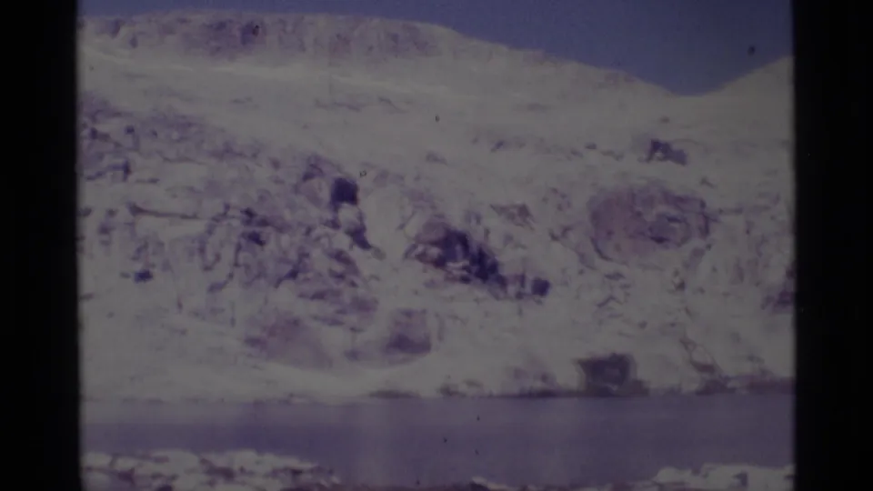 1975 8mm Minaret Wilderness view of a snow-dusted rocky slope above a calm lake, framed by dark film edges.