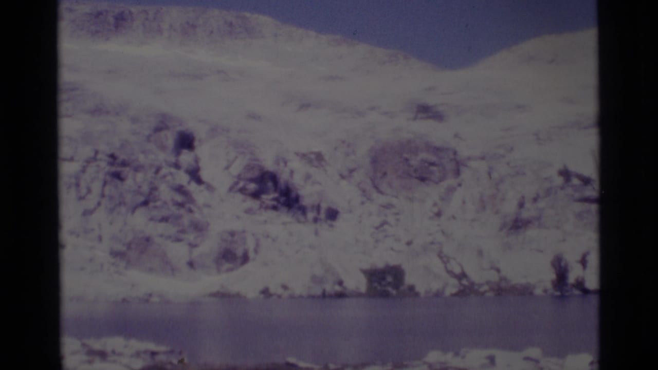 1975 8mm view of a snow-covered Minaret Wilderness mountain slope rising beyond a still lake.