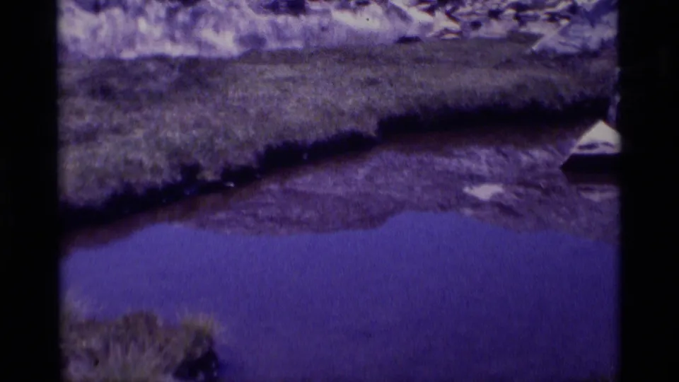 1975 8mm home-movie frame from Minaret Wilderness showing purple pond water, grassy bank, rocks, and a small triangular object near the edge.
