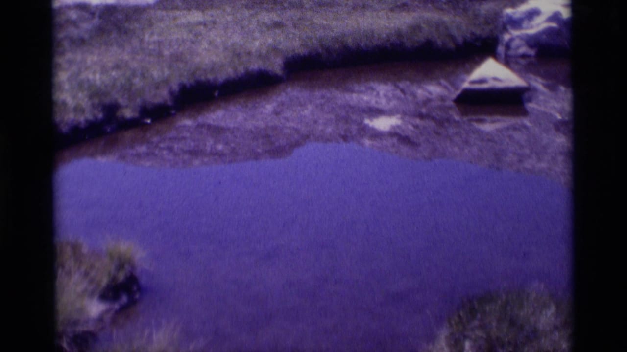 1975 8mm frame from Minaret Wilderness with purple-toned pond water, grassy shore, rocky ground, and a small triangular floating object.