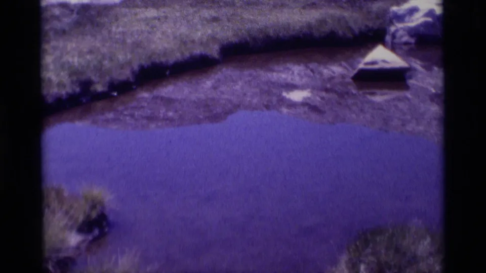1975 8mm frame from Minaret Wilderness with purple-toned pond water, grassy shore, rocky ground, and a small triangular floating object.