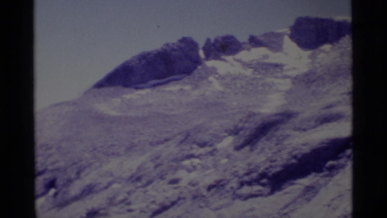 Rocky mountain slope with patches of snow and dark outcrops under a pale sky.