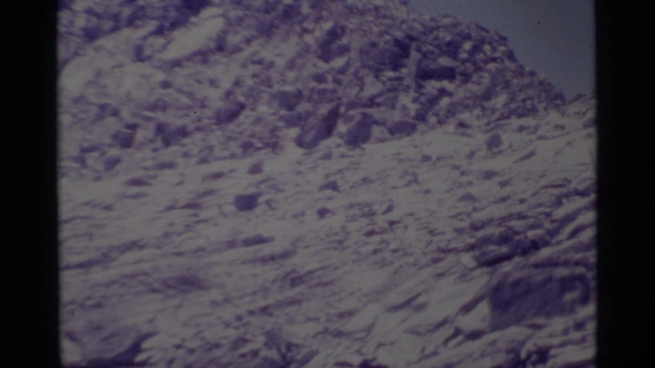 1975 Minaret Wilderness home-movie frame showing a steep rocky slope with patches of snow and a washed pale sky.