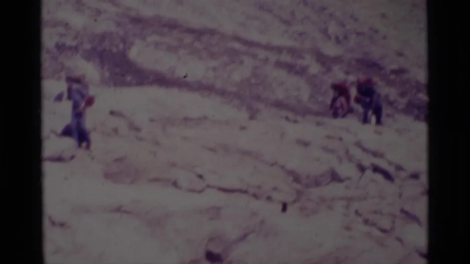 Several distant hikers in dark outdoor clothing cross a snow-dusted slope in 1975 Minaret Wilderness home-movie footage.