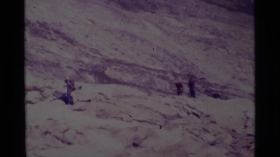 1975 8mm frame from Minaret Wilderness showing small hikers moving across a sunlit rocky slope in a dry mountain landscape.