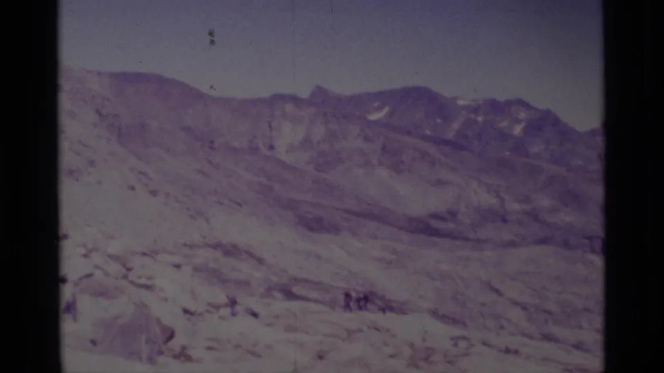 1975 8mm frame from Minaret Wilderness showing distant hikers at the edge of a broad rocky basin and mountain ridge view.
