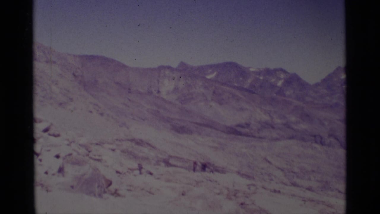 Snow-covered mountain slope and ridgeline with tiny distant figures below a pale sky.