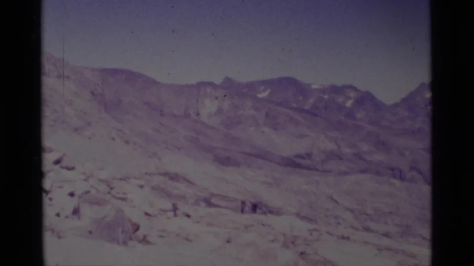 1975 8mm home-movie frame of Minaret Wilderness snowfields, rocky ridges, and tiny distant people in the lower landscape.
