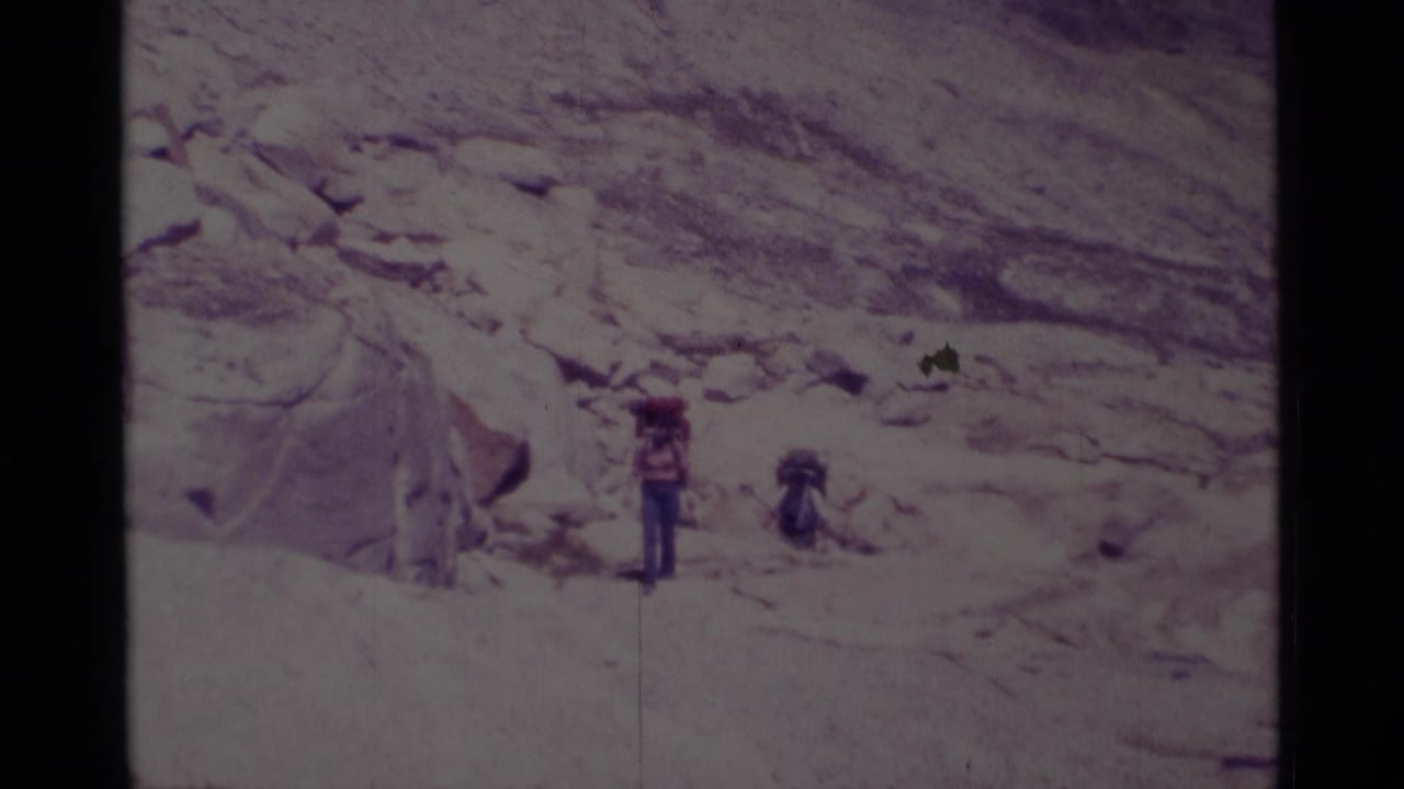 1975 Minaret Wilderness home-movie frame showing two hikers with large packs near granite boulders on a rocky trail.