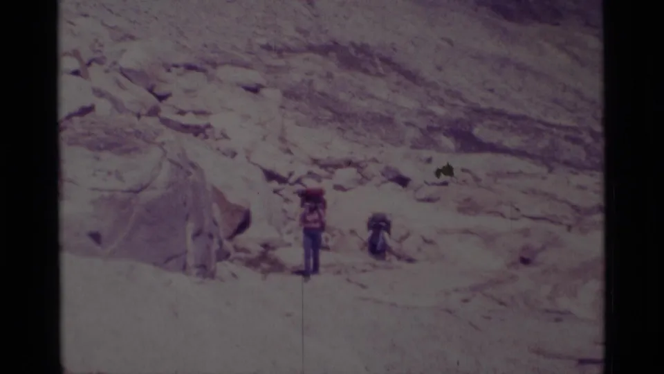 1975 Minaret Wilderness home-movie frame showing two hikers with large packs near granite boulders on a rocky trail.