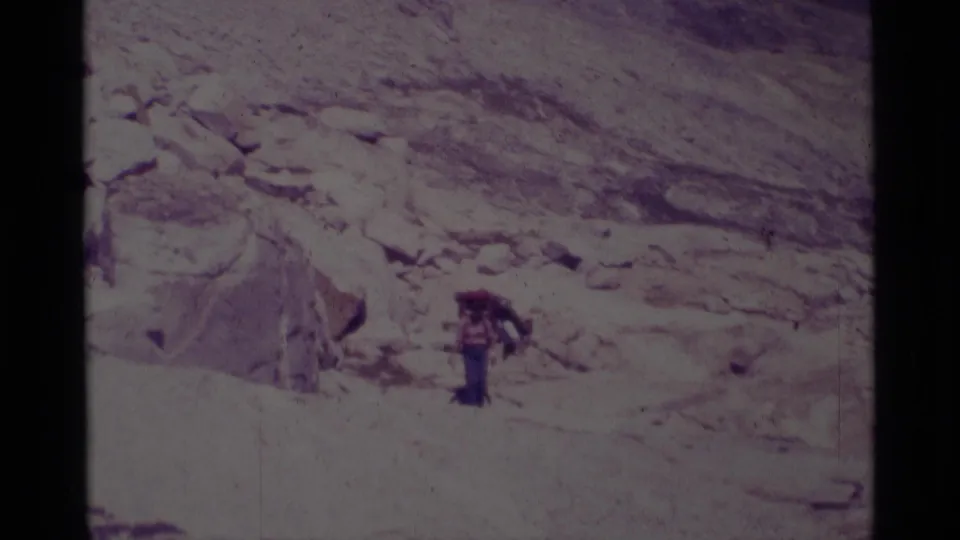 A single visible backpacker stands near pale boulders in 1975 California wilderness footage, with another figure partly obscured nearby.