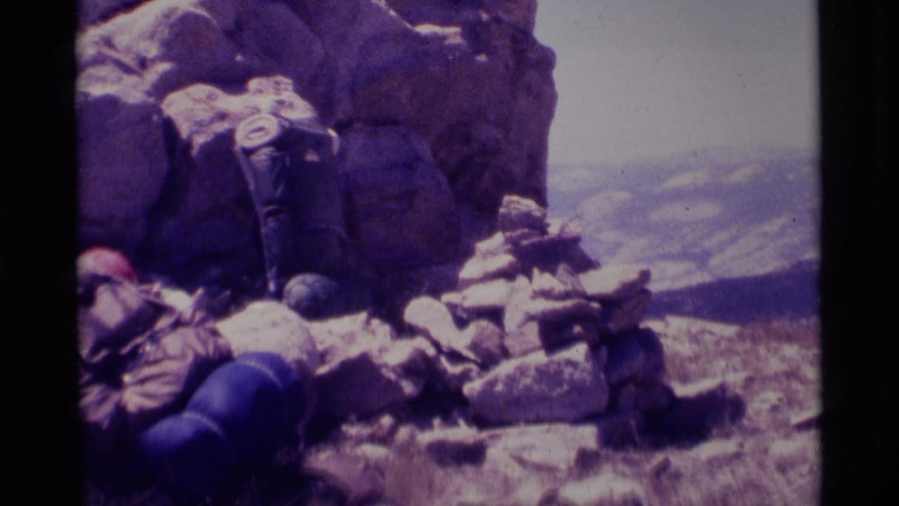 Hikers in a 1975 California wilderness home movie rest below large rocks with backpacks and a hazy valley beyond.