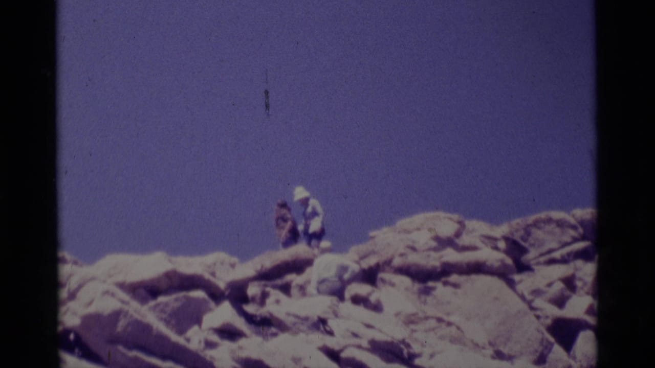 1975 Minaret Wilderness home-movie frame showing a distant hiker on a rocky alpine ridge under a blue sky.