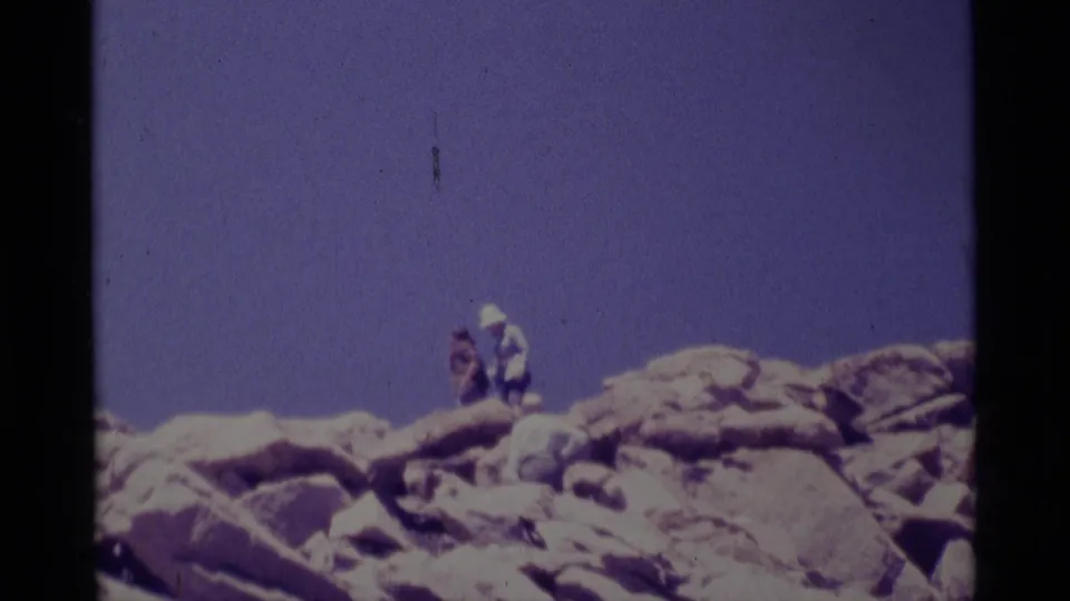 1975 Minaret Wilderness home-movie frame showing a distant hiker on a rocky alpine ridge under a blue sky.