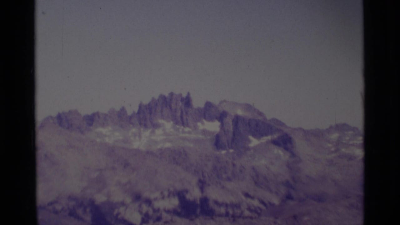 A 1975 home-movie frame shows the Minaret Wilderness landscape from a distance, with sharp rocky spires and snow remnants softened by 8mm color shift.