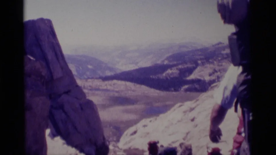 A backpacked hiker stands near rocks in the 1975 Minaret Wilderness footage, looking across snowy slopes and distant mountains.