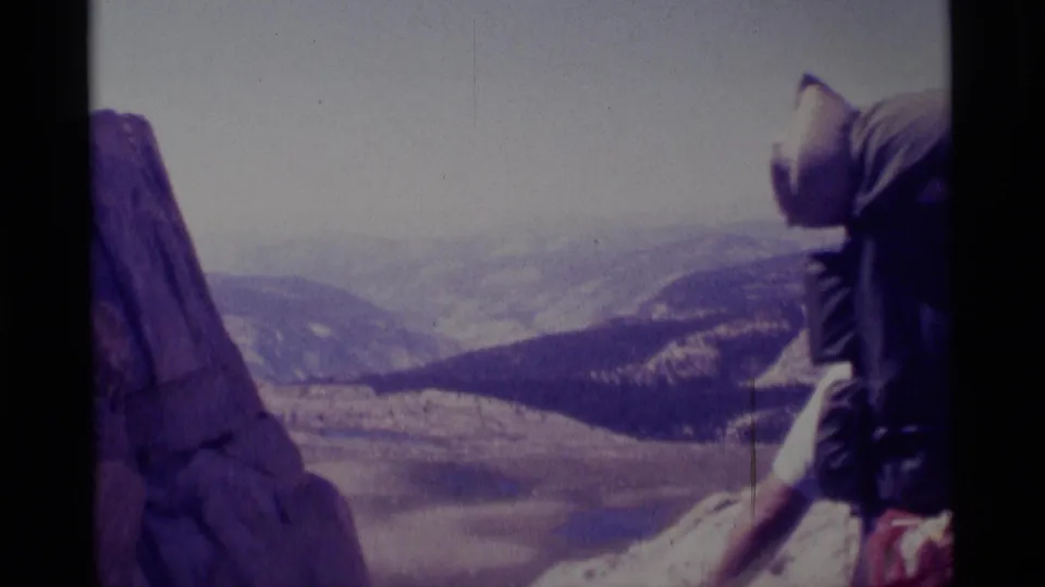 1975 8mm frame from Minaret Wilderness showing a hiker with a large pack near rock above a distant mountain valley.