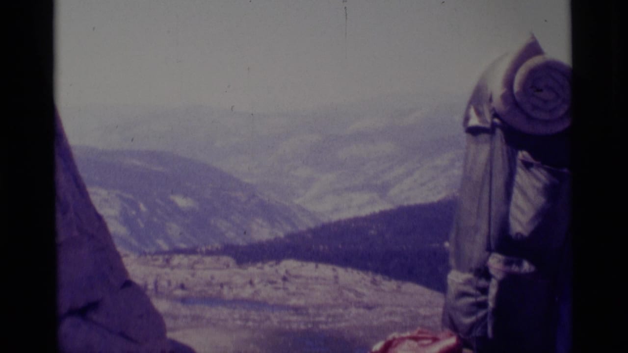 1975 Minaret Wilderness home-movie frame with a backpacked hiker at a rocky high point and mountains beyond.
