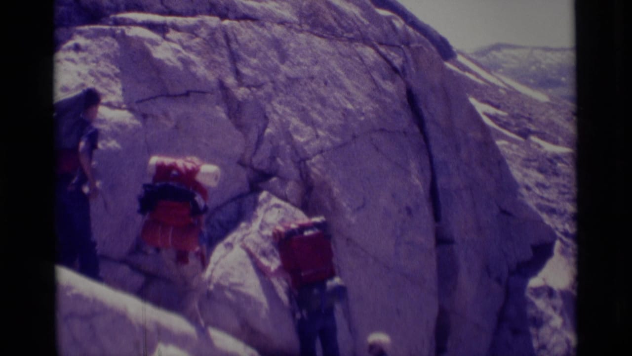 1975 Minaret Wilderness home-movie frame showing hikers with red backpacks moving along a steep rocky slope beside a pale rock wall.