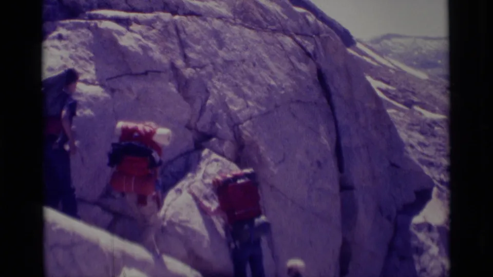 1975 Minaret Wilderness home-movie frame showing hikers with red backpacks moving along a steep rocky slope beside a pale rock wall.