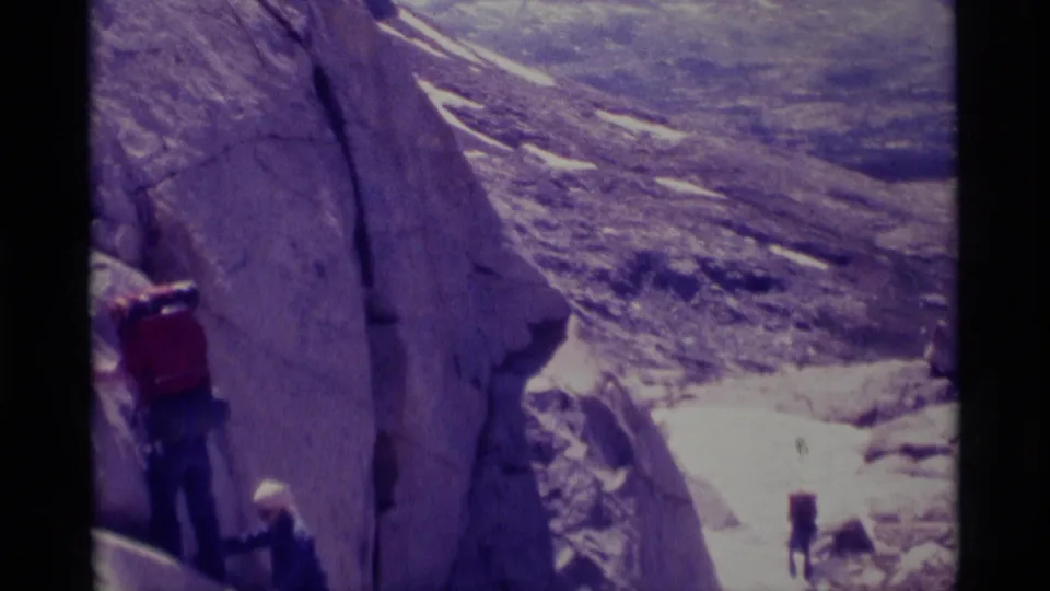A hiker carrying a red backpack moves near a pale rock wall while another person stands lower on the slope in the 1975 wilderness scene.