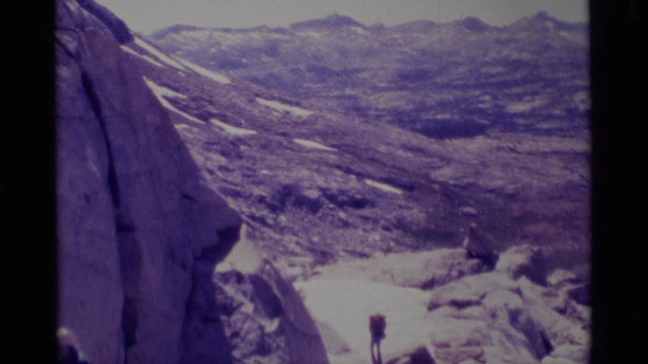 1975 Minaret Wilderness home-movie frame showing pale granite, snow patches, distant ridgelines, and a small hiker for scale.
