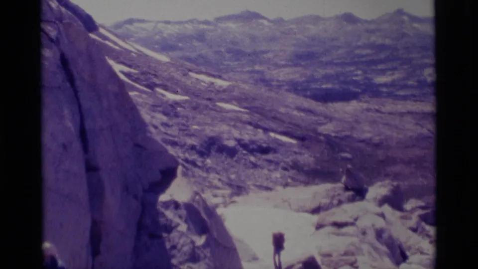1975 Minaret Wilderness home-movie frame showing pale granite, snow patches, distant ridgelines, and a small hiker for scale.