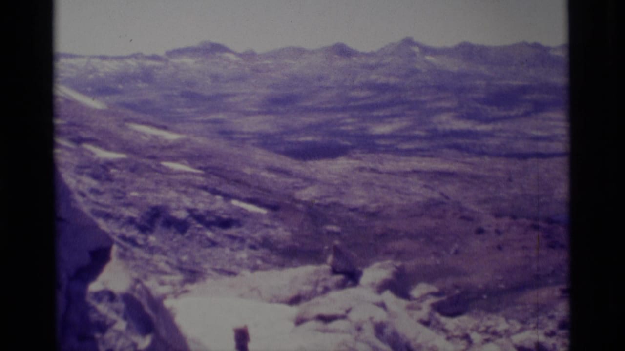 Wide 1975 California wilderness view with rocky terrain, lingering snow patches, distant ridgelines, and a small hiker partly visible.