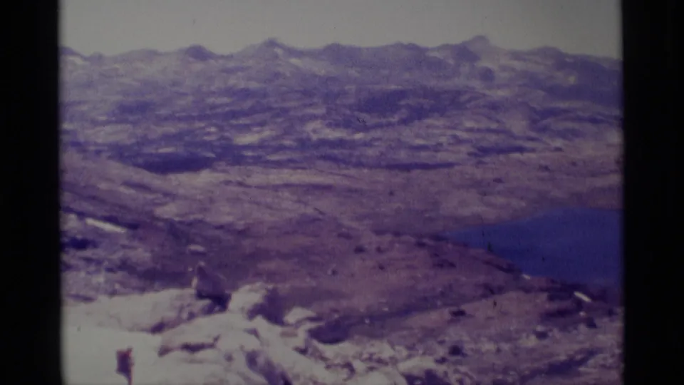 Minaret Wilderness frame from 1975 showing rugged slopes, scattered snow, a distant blue alpine lake, and soft 8mm color.