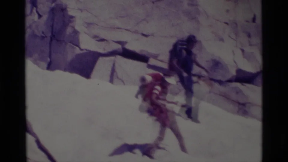 Two distant climbers traverse a steep rock face in 1975 Minaret Wilderness footage, with one person wearing a red backpack.