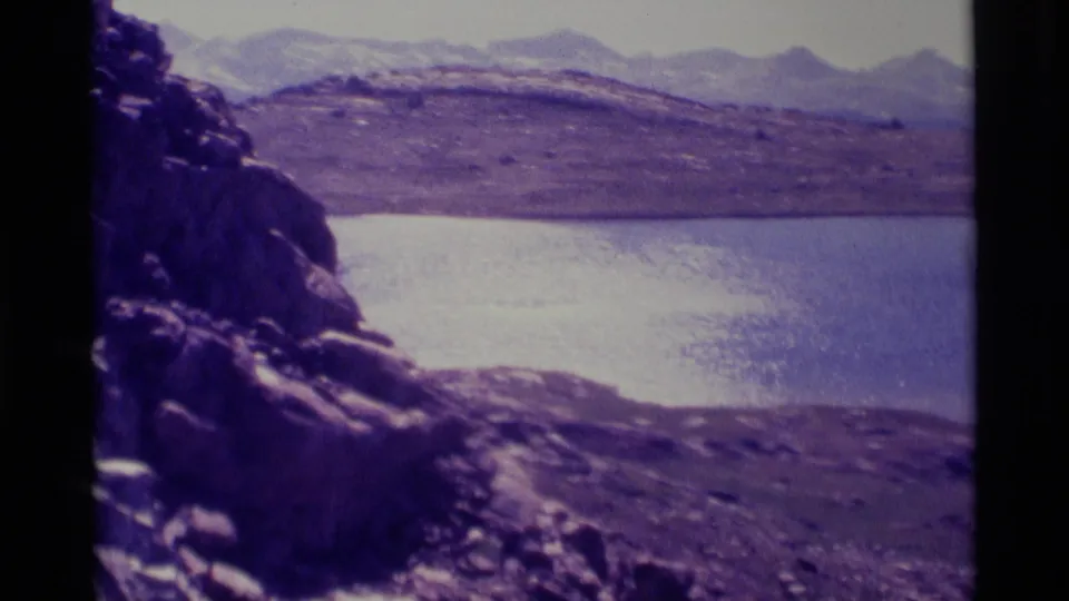1975 8mm frame showing a calm alpine lake, rocky foreground, open slopes, and distant ridges near Minaret Wilderness.