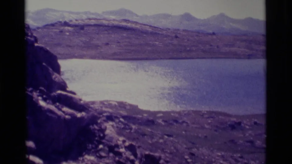 Final 8mm frame of a still mountain lake with rocky shoreline, open hills, and distant peaks in the 1975 California wilderness clip.