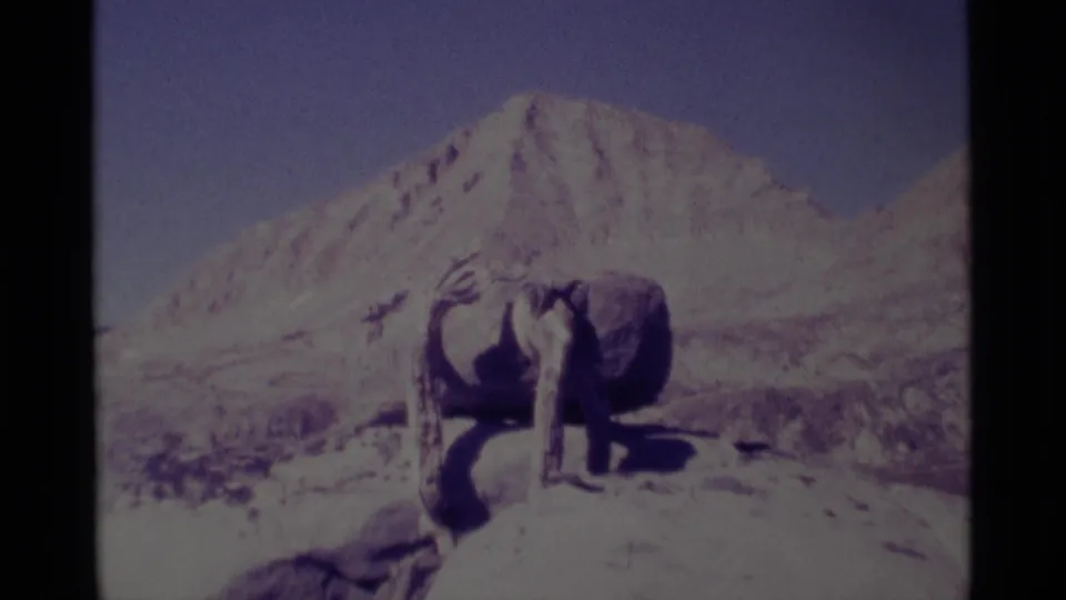 Faded 8mm frame from 1975 showing the abstract sculpture more prominently, with rocky ground and Minaret Wilderness mountains behind.