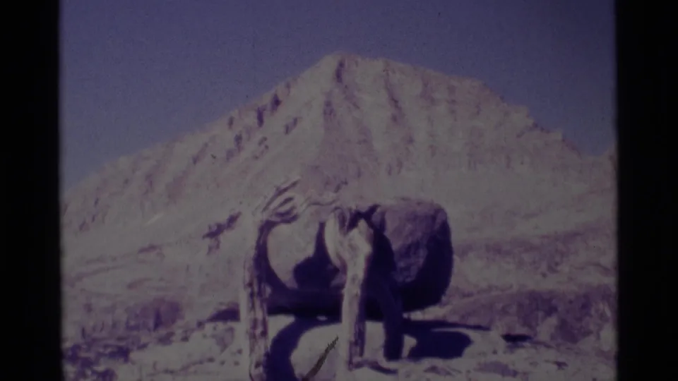 1975 8mm Minaret Wilderness frame showing the sculpture on a pale rock ledge with a large mountain peak and scrubby terrain beyond.