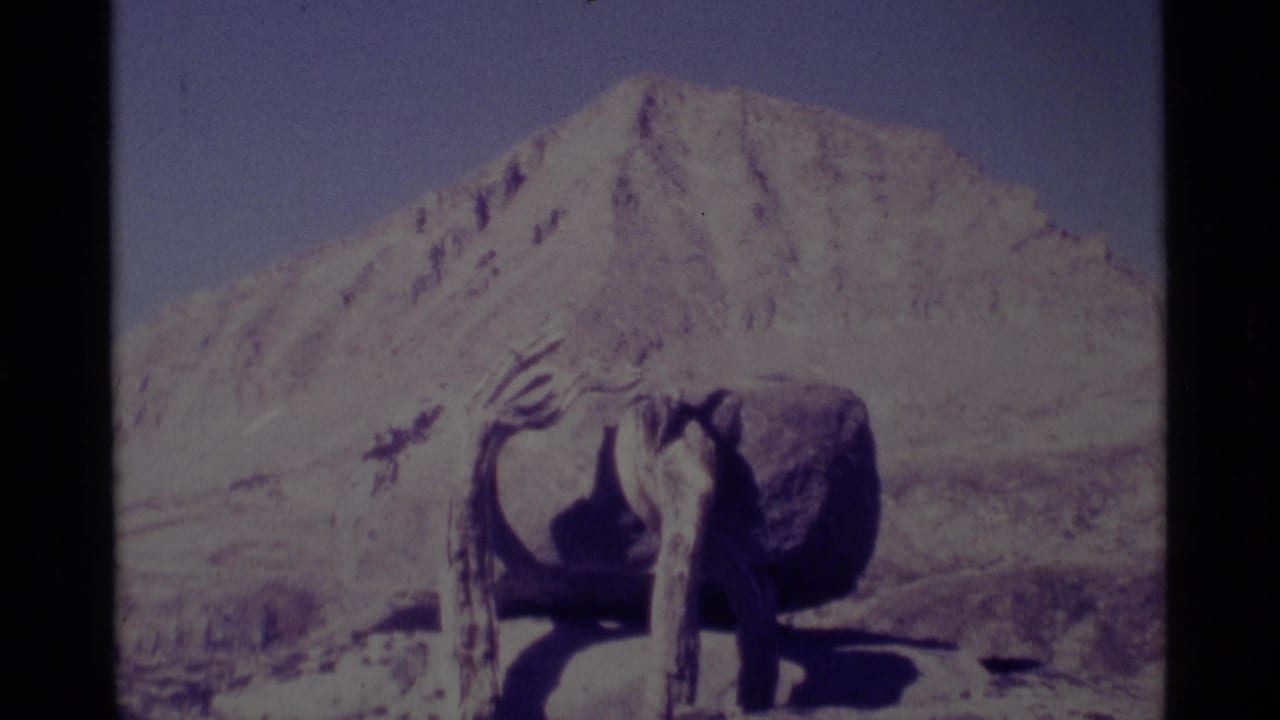 1975 8mm frame from Minaret Wilderness showing a rough wooden structure before a sunlit mountain peak and dry alpine ground.