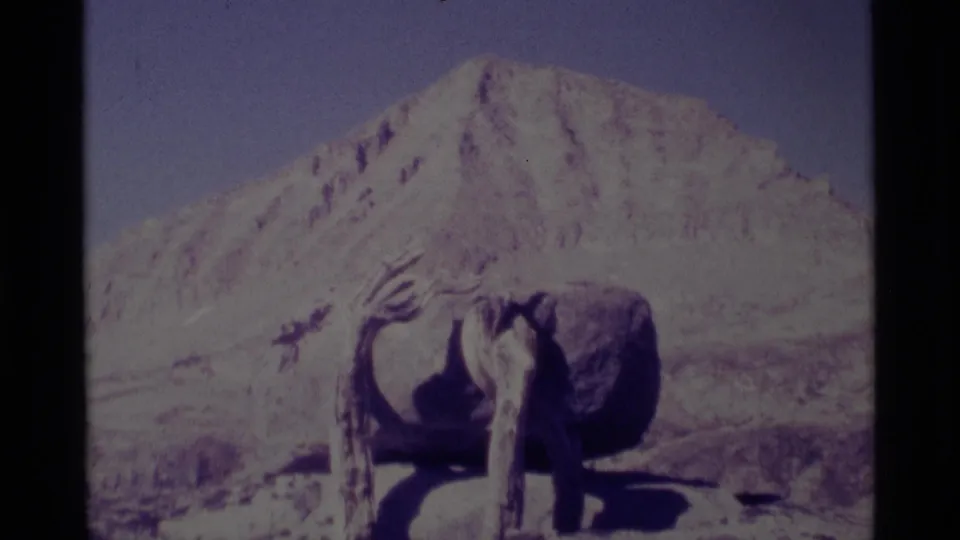 1975 8mm frame from Minaret Wilderness showing a rough wooden structure before a sunlit mountain peak and dry alpine ground.