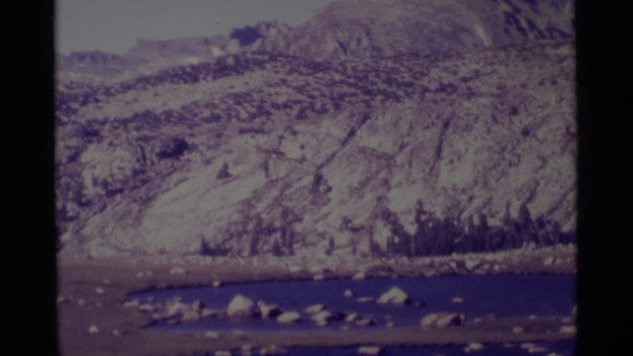 1975 8mm view of a rocky Minaret Wilderness lakeshore with mountain slopes and dark film gate borders.