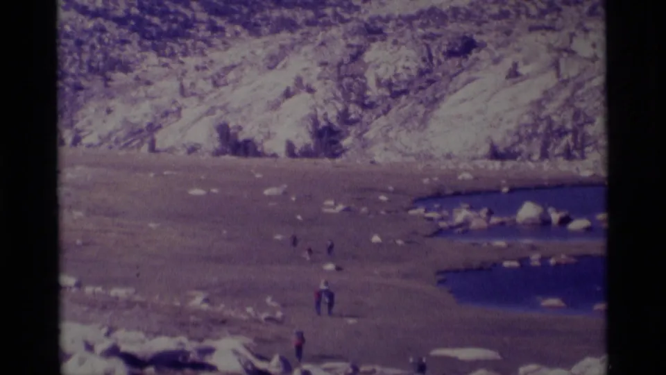 Several distant hikers stand or walk across a rocky Minaret Wilderness lakeshore in faded 1975 8mm color.