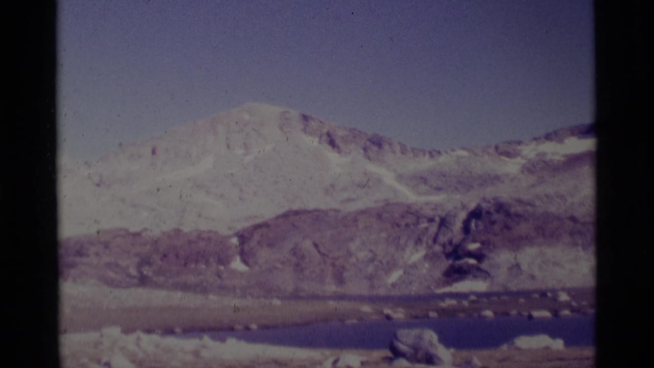 1975 8mm frame of the Minaret Wilderness area showing a snow-dusted ridge, rocky shore, and calm alpine lake under a pale sky.